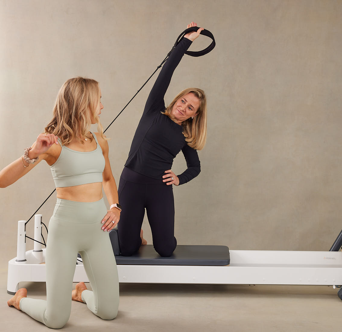 Sam Deville coaches a female client in a Pilates studio using reformer equipment. Sam demonstrates the correct positioning, kneeling with one hand on her hip and looking back at the client. The client mirrors the pose, kneeling with one hand on her hip and the other arm extended overhead, holding a black loop band while stretching to the right.